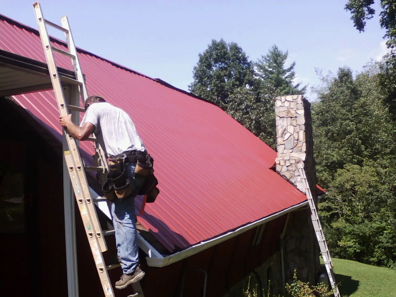 Roofer on a ladder beside a red metal roof and stone chimney — Soffit & Fascia Repair by Roofing Brothers in Mattawana